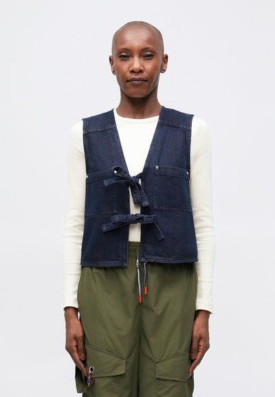 Denim vest in donkerblauw met een striksluiting, twee voorzakken en naadloze schouders, gedragen over een witte langarmshirt.
