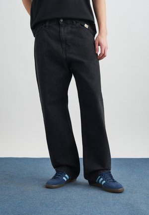 Persona che indossa jeans neri larghi e sneakers blu con strisce azzurre, in piedi su un tappeto blu davanti a uno sfondo chiaro.