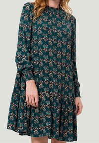 Vestido de un verde azulado oscuro con un patrón floral en rosa y verde, de mangas largas, escote con pliegues y dobladillo con volantes en capas. Textura de tela suave.