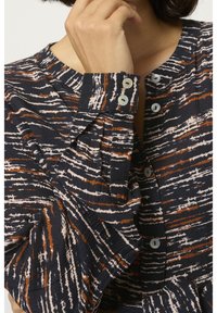 Main de femme près du visage portant une chemise à manches longues avec des rayures abstraites noires, blanches et marron et des boutons blancs.