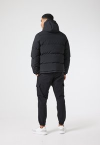 Melnā pūkā jaka ar kapuci, ar quilted dizainu un elastīgiem piedurknēm, apvienota ar melnām kravas biksēm un baltām sporta kurpēm.