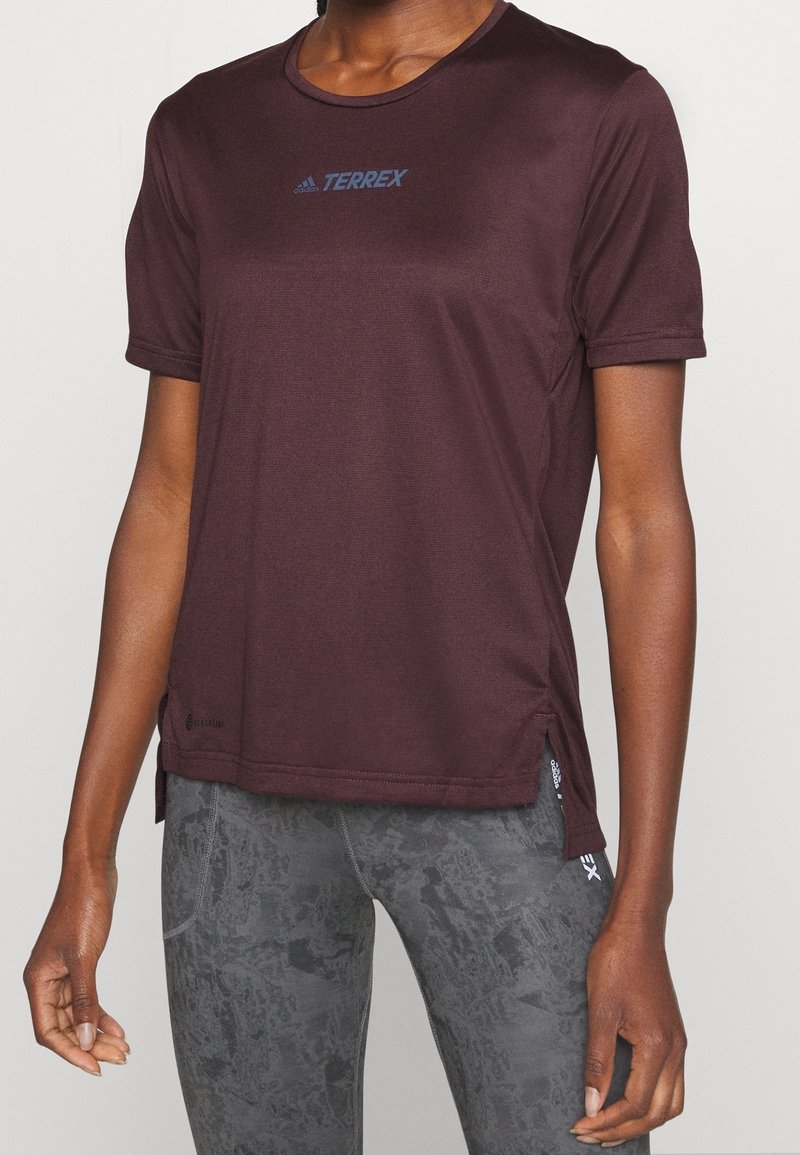 Camiseta deportiva burdeos de manga corta, cuello redondo y aberturas laterales. Presenta un logotipo azul de Adidas Terrex en el pecho.