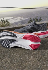 Chaussures de course grises et noires avec une semelle rose, orange et noire, posées sur un pavé rugueux avec un paysage urbain flou en arrière-plan.