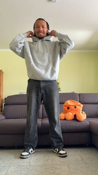 Gray hoodie with drawstrings, loose fit, paired with dark jeans. Black and white sneakers. A plush orange octopus sits on a couch in the background.
