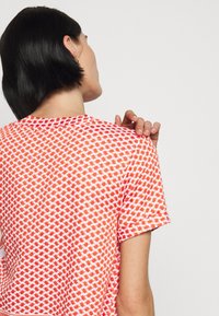 Žena s krátkými tmavými vlasy, oblečená v červeno-bíle vzorované košili s krátkým rukávem, dotýká se pravého ramena levou rukou.