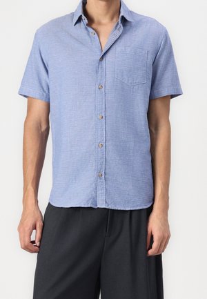 Homme portant une chemise à manches courtes bleu clair texturée avec poche poitrine et un pantalon foncé ample sur un fond uni.