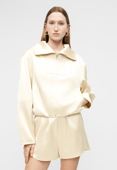 Γυναίκα με μακριά μαλλιά που φοράει μια κρεμ ζακέτα με φερμουάρ και μεγάλο γιακά, καθώς και σορτς στο ίδιο χρώμα, που στέκεται μπροστά σε απλό ανοιχτόχρωμο φόντο.