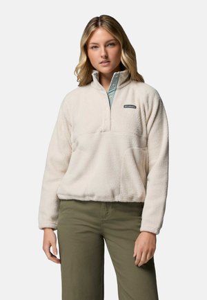 Woman wearing a cream fleece pullover with a stand collar and snap buttons paired with olive green pants, standing against a plain background.