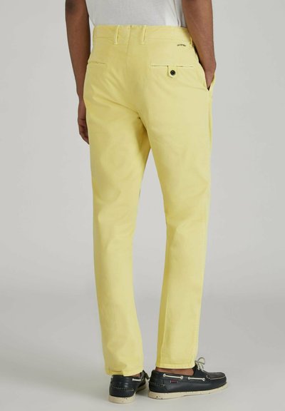 Pantaloni in cotone giallo con taglio dritto, due tasche posteriori e un dettaglio con bottone. Abbinati a scarpe scure e cuciture visibili.