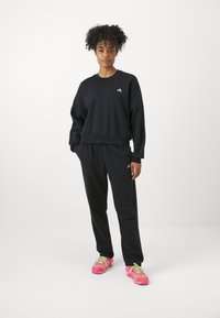 Μαύρη προπόνηση και παντελόνι φόρμας σεσέτ, με άνετη εφαρμογή, ριμπ μανσέτες και διακριτό λογότυπο. Το μοντέλο φοράει ζωντανά ροζ αθλητικά παπούτσια.