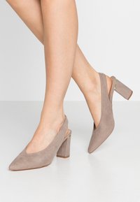 Kvinna som bär sandfärgade mocka slingback blockklackskor med spetsiga tår, stående på en ljus golv mot en enfärgad bakgrund.