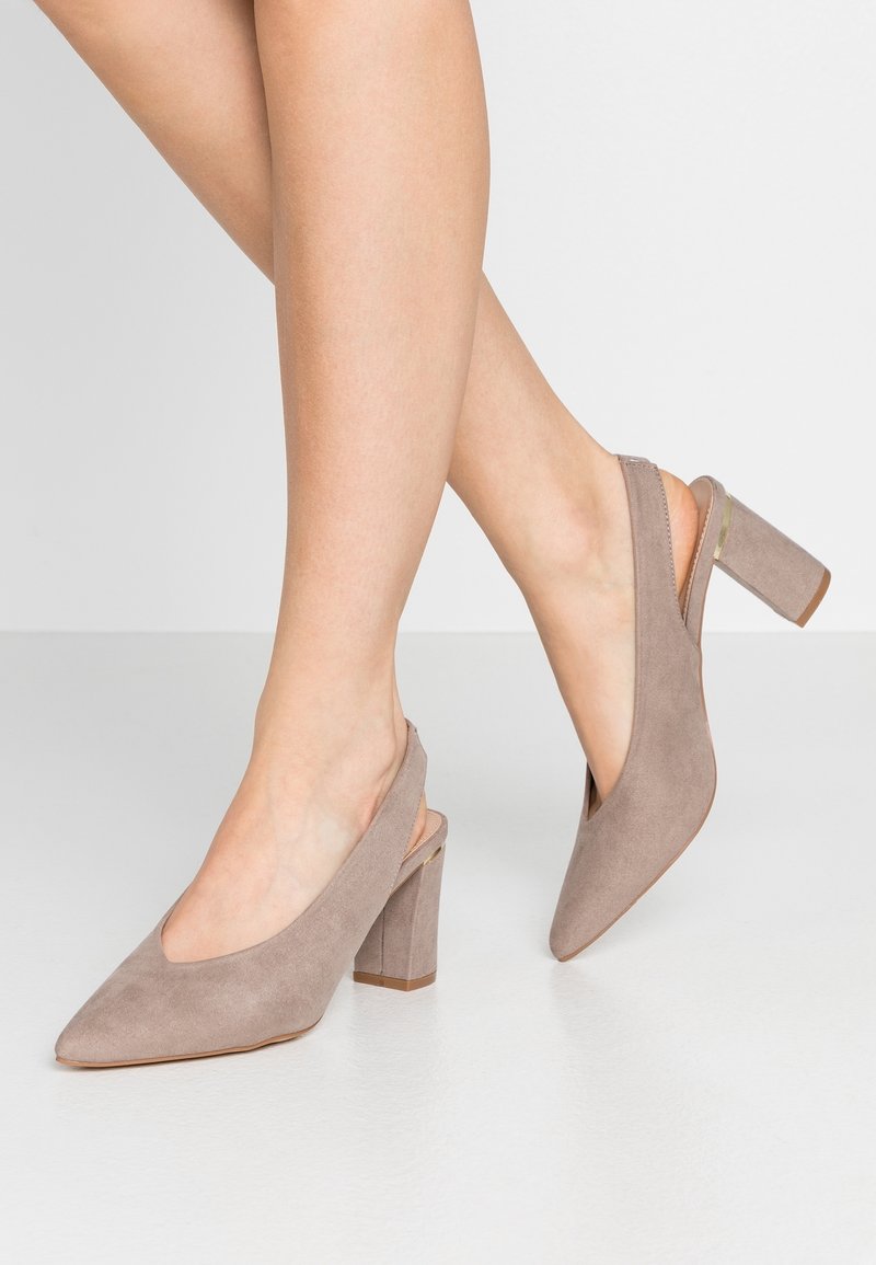 Kvinna som bär sandfärgade mocka slingback blockklackskor med spetsiga tår, stående på en ljus golv mot en enfärgad bakgrund.