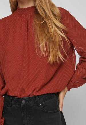 Kvinde iført rust-rød struktureret bluse med lange ærmer stoppet ned i sort høj-taljede jeans, hånd hvilende på hoften, med langt blond hår.