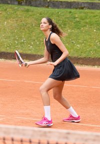 Jogadora de ténis feminina vestida de preto e com sapatos cor-de-rosa prepara-se para bater na bola num court de terra batida com fundo de relva verde.