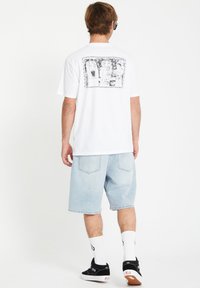 T-shirt blanc à manches courtes avec un design graphique noir au dos, associé à un short en jean bleu clair et des baskets noires.
