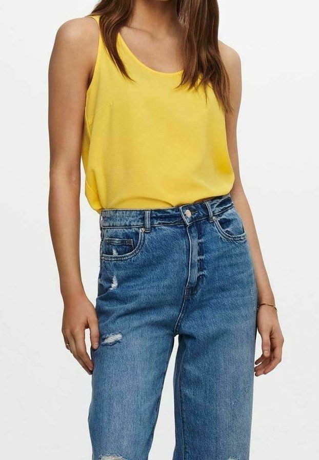 Femme portant un haut jaune sans manches et un jean bleu taille haute légèrement usé, debout devant un fond blanc uni.