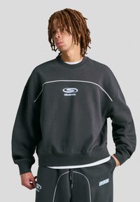 Sweatshirt nero con cuciture e logo bianchi. Presenta un collo girocollo e una vestibilità oversize. Indossato con pantaloni della tuta neri abbinati.