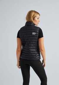 Schwarze gepolsterte Weste mit hohem Kragen, versehen mit horizontalen Nähten und einem Logo-Detail auf dem Rücken, getragen über enganliegenden schwarzen Leggings.