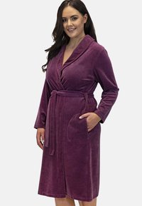Accappatoio viola in peluche con maniche lunghe, colletto a scialle, vita con cordoncino e due tasche frontali. Texture morbida e design fino al ginocchio.