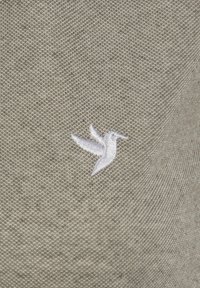 Grau gestrickter Stoff mit einem kleinen, weißen gestickten Kolibri-Logo, das mit sichtbarer Textur und einem einzigartigen Design versehen ist.