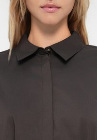 Detailní záběr ženy, která nosí černou blůzku s límečkem a skrytým zapínáním, a má na rtech lesklou světle růžovou rtěnku.