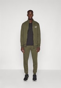 Survêtement vert olive en matériau doux, comprenant une veste zippée avec un logo et des poignets élastiques, assorti de joggers et de baskets noires.