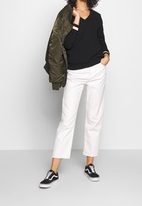 Czarny sweter z dekoltem w serek, oliwkowa kurtka typu bomber, białe spodnie o kroju 7/8 oraz czarne trampki z białymi sznurowadłami. Codzienny strój z kontrastującymi kolorami.