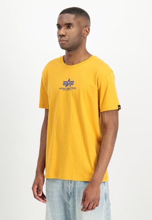 Mann trägt ein gelbes Alpha Industries T-Shirt mit blauem Logo, steht vor einem schlichten weißen Hintergrund und schaut leicht nach links.