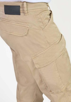 Beige cargo pants with multiple pockets, including side flap pockets and a small front coin pocket, worn with a tucked-in white shirt.