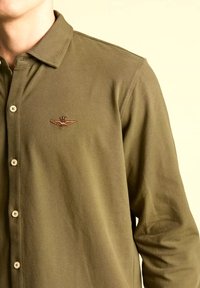 Camicia verde oliva a maniche lunghe con colletto, caratterizzata da un piccolo stemma ricamato con ali e bottoni di colore chiaro. Tessuto di cotone liscio.