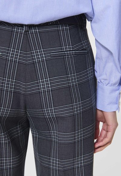 Czarno-szare spodnie w kratę wykonane z teksturowanej tkaniny, mające dopasowany krój i kieszenie z tyłu. Model ma na sobie jasnoniebieską koszulę.