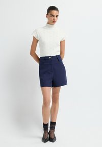 Vrouw in een wit gebreid T-shirt met korte mouwen, marineblauwe high-waisted shorts met flapzakken, doorzichtige zwarte sokken en zwarte puntige hakken, staande.