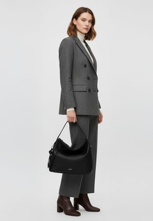 Woman in grey double-breasted suit and brown ankle boots holds large black leather handbag against plain background.