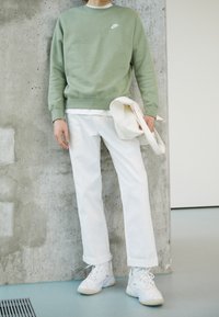 Sudadera verde con logo blanco, camiseta blanca debajo, pantalones rectos blancos y zapatillas blancas con suelas texturizadas, de pie contra una pared de hormigón.
