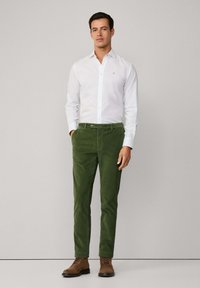 Man in een witte lange mouwen overhemd, olijfgroene corduroy broek en bruine veterschoenen, staand tegen een effen achtergrond.