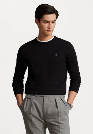 Sort crewneck sweater med ribbede manchetter, parret med lysegrå plisserede bukser. Subtil logo detalje på brystet. Glat tekstur.