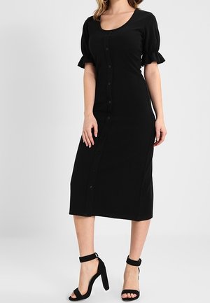 Strak zwarte midi-jurk met knopen aan de voorzijde, korte pofmouwen en ronde hals, gedragen met zwarte enkelsandaaltjes met hoge hakken.