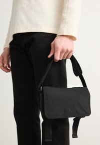 Svart nylon skulderveske med rektangulær form, med justerbare stropper og en logo preget på fronten. Jevn tekstur.