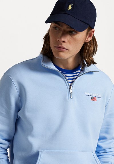 Jasnoniebieski polar z półzamkiem, z logo na przodzie i kieszenią kangurką. Noszony na paskowanym niebieskim t-shircie.