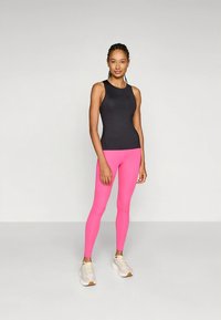 Μαύρο τοπ χωρίς μανίκια και φωτεινό ροζ κολάν, με λεία υφή. Τα αθλητικά παπούτσια είναι ανοιχτό γκρι με λευκές και καφέ λεπτομέρειες.