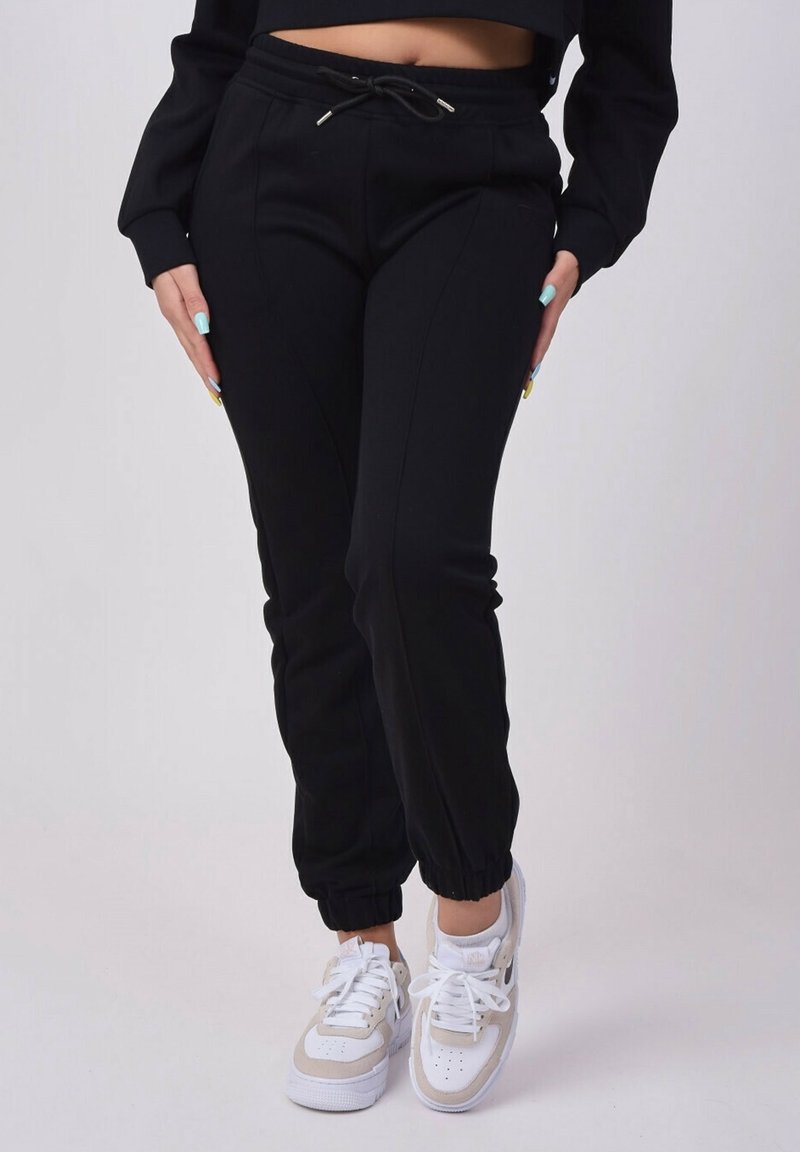 Osoba ubrana w czarne dresowe spodnie i białe sneakersy, stojąca z rękoma opartymi na biodrach na jednolitym tle.