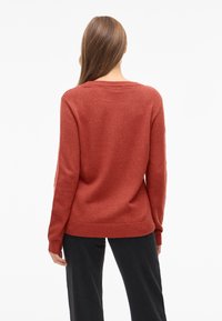 Kvinne med langt brunt hår iført en rustfarget genser og svarte bukser, stående vendt bort fra kameraet mot en hvit bakgrunn.