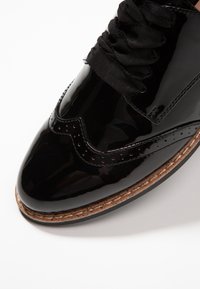 Chaussure en cuir verni noir avec une finition lisse et brillante, agrémentée d'accents perforés et de lacets noirs, avec une semelle à texture liège.