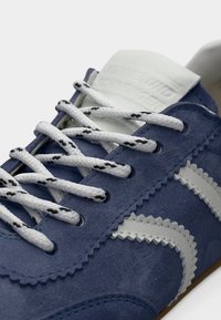 Κοντινό πλάνο ενός μπλε σουέντ αθλητικού παπουτσιού με λευκές ζιγκ-ζαγκ λεπτομέρειες και λευκά κορδόνια με μαύρα πουά.