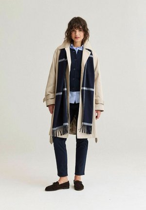 Kvinde iført beige trenchcoat med marineblåt ternet halstørklæde, marineblå cardigan, stribet skjorte, mørke bukser og mokkassinsko, stående mod en ensfarvet baggrund.