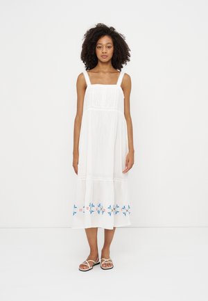 Woman with curly hair wearing a white sleeveless midi dress with blue and pink floral embroidery near the hem and white sandals.