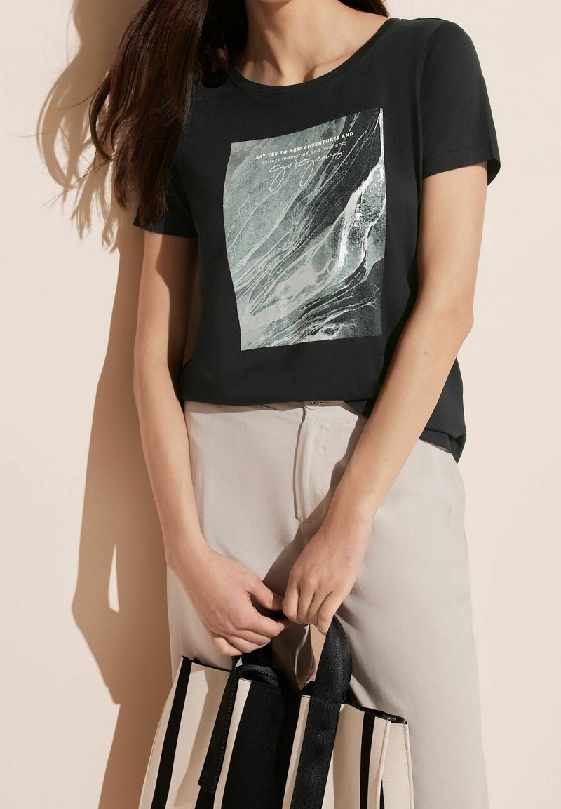 Žena v černém tričku s abstraktním potiskem a textem, v béžových kalhotách, držící černobílou pruhovanou tašku před světlým pozadím.