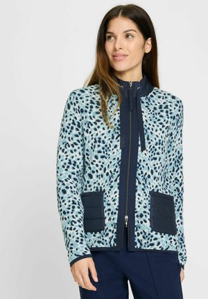 Veste bleu clair avec imprimé léopard noir, fermeture éclair à l'avant, accents navy et deux poches matelassées. Dotée d'un col haut et de manches longues.
