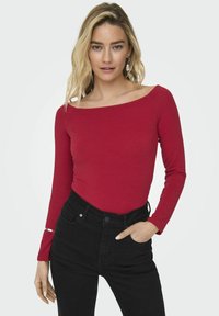 Crvena majica dugih rukava s brojnim izrezom, koja se ističe uskim rezom. Uparena s crnim trapericama visokog struka. Glatka tekstura, minimalni detalji.