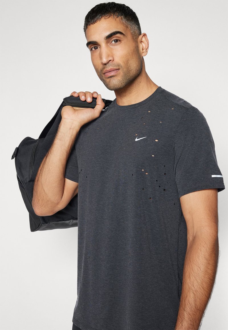 Maglietta Nike grigia scura con multiple perforazioni, maniche corte e logo sul petto. Modello che tiene una borsa da palestra nera.